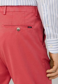 Rode stoffen broek met een achterzak, voorzien van een knoop en een label met "HACKETT." Gecombineerd met een lichtblauwe gestreepte overhemdsmouw.