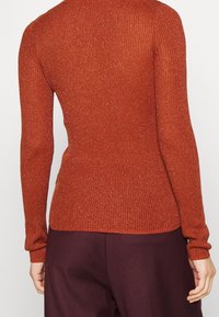 Vue arrière d'une personne portant un pull orange ajusté à manches longues en côte et un pantalon violet foncé, se tenant devant un fond uni.