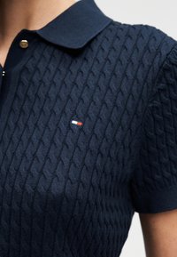 Gros plan d’un polo en maille bleu marine à manches courtes, texture à motif câblé et petit logo rectangulaire rouge, blanc et bleu sur la poitrine.