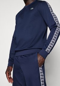 Námořnický sportovní svetr z polyesteru s dlouhými rukávy, vroubkovanými manžetami a grafickým lemem po stranách. Na hrudi má malé zelené logo.