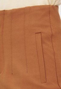 Gros plan sur la ceinture d'une jupe couleur camel avec des coutures verticales et une poche fendue, portée avec un haut côtelé crème.