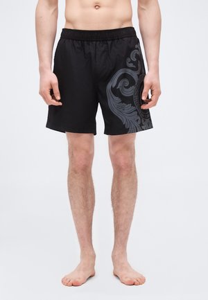 Uomo che indossa pantaloncini da bagno neri con motivo barocco grigio scuro sulla gamba destra, in piedi a piedi nudi su uno sfondo bianco uniforme.