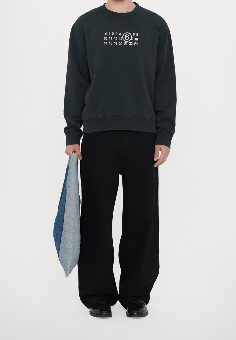 Personne portant un sweat-shirt noir avec des chiffres blancs de calendrier et un 6 encerclé, pantalon large noir, chaussures noires, tenant un tissu bleu plié dans la main gauche.
