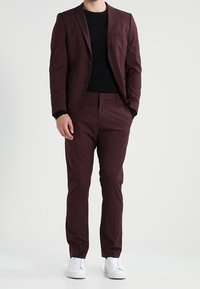 Costume bordeaux avec une coupe ajustée ; veste à simple boutonnage, revers crantés, et pantalon slim, associé à une chemise noire texturée et des baskets blanches.