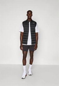 Svart vadderad väst över en vit kortärmad skjorta, kombinerad med grå shorts. Västen har horisontell vaddering och en framdragkedja.