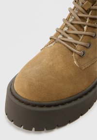 Hnědé semišové kotníkové boty s tlustou černou platformou, s šněrováním a kontrastním prošíváním podél okraje.