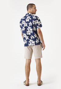 Camisa de manga corta azul marino con patrón floral blanco, combinada con pantalones cortos beige claro y zapatos slip-on marrones, vista desde atrás.