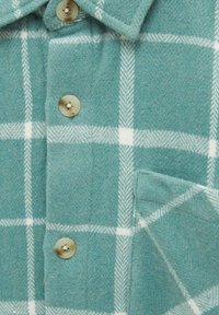 Primo piano di una camicia di flanella color teal con un motivo a quadri bianchi, con bottoni color sabbia e una tasca sul petto. È visibile un tessuto morbido e testurizzato.