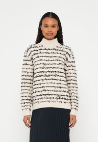Femme portant un pull à col roulé crème avec des rayures horizontales texturées noires et une jupe plissée noire, debout devant un fond blanc.