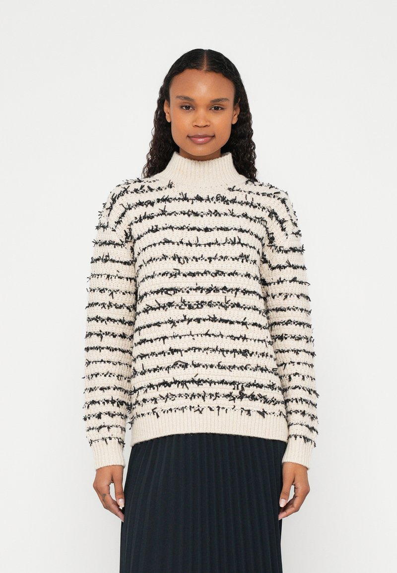 Femme portant un pull à col roulé crème avec des rayures horizontales texturées noires et une jupe plissée noire, debout devant un fond blanc.