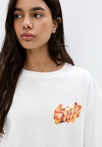 T-shirt bianca in cotone con vestibilità oversized, caratterizzata da un design colorato che riporta la scritta "Sunk" con un accento ciliegia nella zona del petto sinistro.