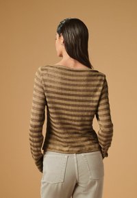 Brunstribet langærmet bluse med en bred udskæring og tekstureret stof, parret med lyse beige bukser. Neutral baggrund.