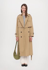 Cappotto beige con design doppiopetto, ampie tasche e spalline, abbinato a pantaloni color crema e ballerine nere, mentre tiene una borsa verde.