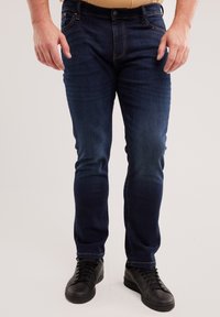 Muškarac u tamnoplavim uskim trapericama i crnim tenisicama s vezicama stoji nasuprot jednobojnoj svijetloj pozadini.