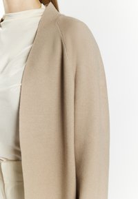 Beige cardigan van zacht materiaal, met een losse pasvorm, verlaagde schouders en een open voorkant. De gladde textuur contrasteert met het lichte shirt.