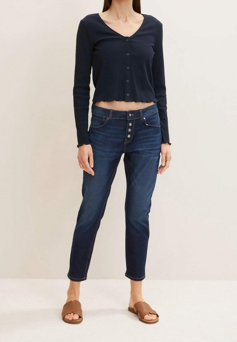 Mujer que lleva un cárdigan corto abotonado de color azul oscuro, unos jeans de tiro alto azul oscuro con múltiples botones y unas sandalias de deslizar marrón.