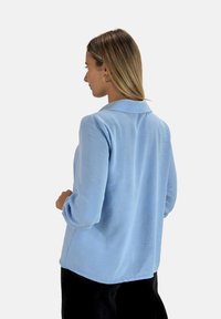 Chemise bleu clair à manches longues avec un col et un tissu texturé, dotée d'une coupe décontractée et d'un ourlet légèrement arrondi.