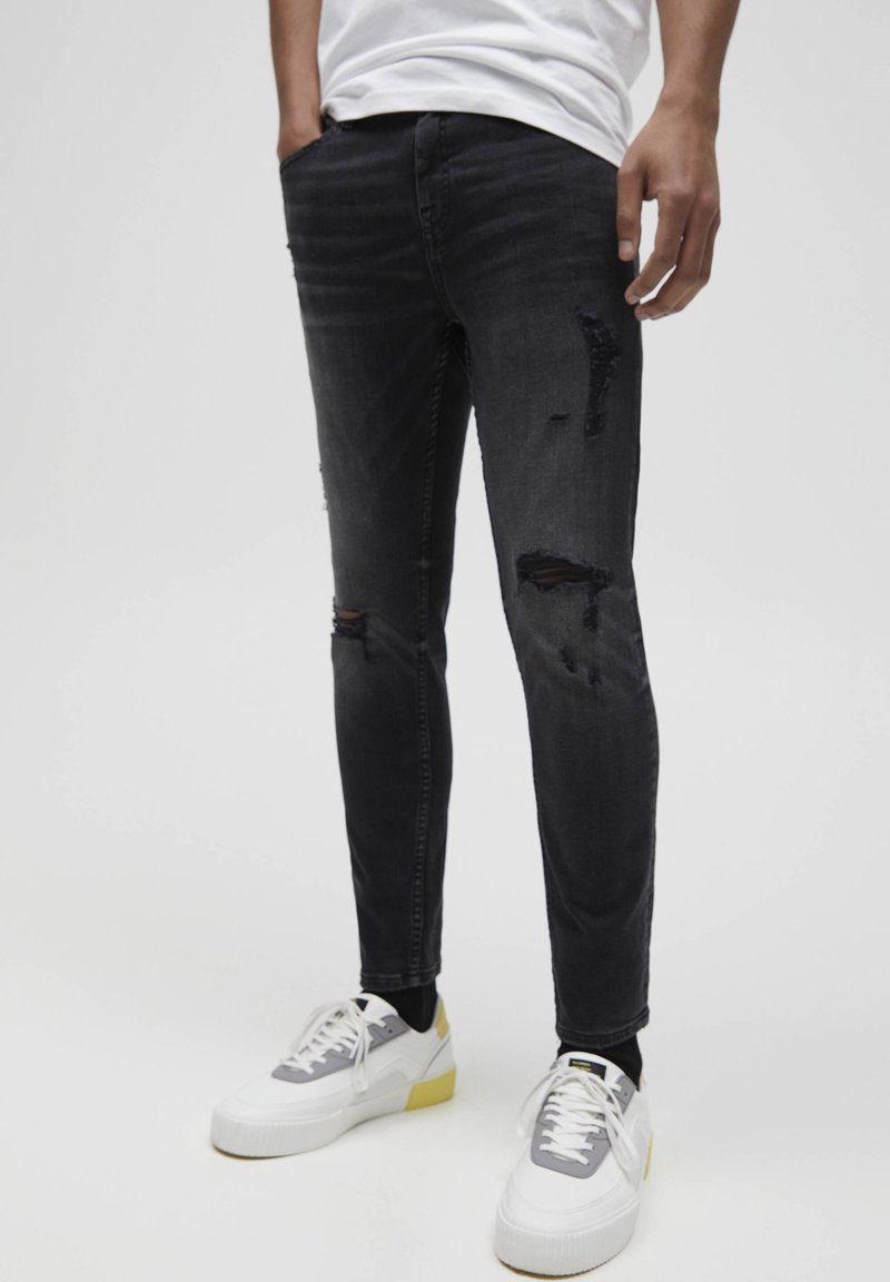 Person klädd i svarta trasiga skinny jeans, vita sneakers med grå och gula detaljer, och en vit skjorta, stående mot en neutral bakgrund.