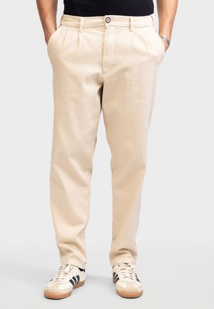 Beige broek met plooien, gemaakt van een gladde stof. Voorzien van een knoopsluiting en twee achterzakken, gedragen met crème en zwarte sneakers.