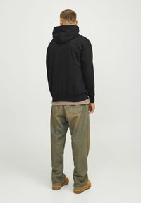 Sudadera negra con capucha y puños acanalados, llevada sobre una camiseta de color beige claro. Jeans desgastados en tonos verde marrón y botas de trabajo beige visibles.