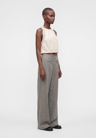 Femme debout de profil portant un haut beige sans manches avec bordure en dentelle et un pantalon gris taille haute à jambes larges avec des chaussures noires.
