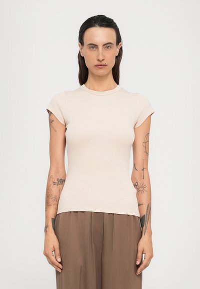 Persona con cabello largo y oscuro y tatuajes en ambos brazos, vistiendo una camiseta de manga corta beige y pantalones sueltos marrones, de pie frente a un fondo liso.