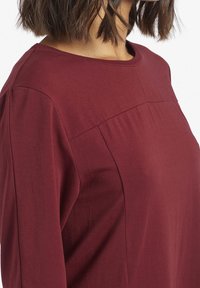 Top bordeaux realizzato in morbido tessuto elasticizzato. Presenta un colletto rotondo, spalle scese e pieghe sottili lungo le cuciture laterali.
