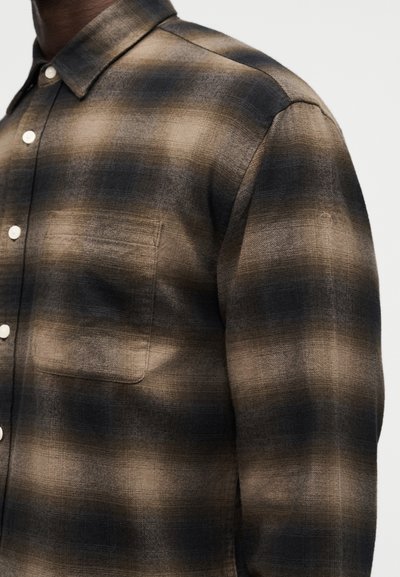 Άνδρας που φοράει πουκάμισο με καρό μοτίβο σε μπεζ και σκούρο καφέ, με τσέπη στο στήθος, εμφανιζόμενος από το μέσο του στήθους έως τον ώμο, μπροστά σε απλό φόντο.
