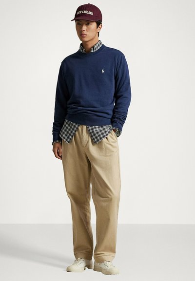 Maglione blu navy girocollo sopra una camicia a quadri, pantaloni beige e scarpe da ginnastica bianche. Cappellino marrone con scritto "NEW ENGLAND" in testo bianco.