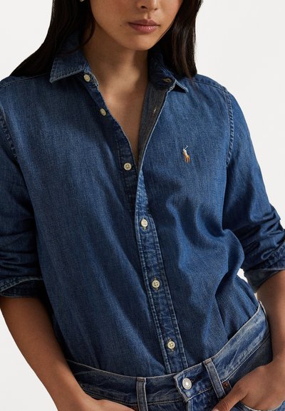 Camisa de ganga azul-escura com botões, apresentando um colarinho pontiagudo, costuras visíveis e um pequeno logo bordado no peito.