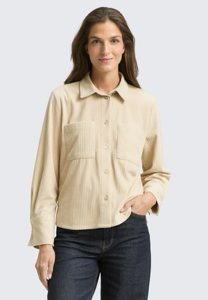 Camicia in corduroy beige con colletto, due tasche frontali e chiusura a bottoni. Maniche lunghe con dettaglio ai polsini. Indossata con jeans scuri.
