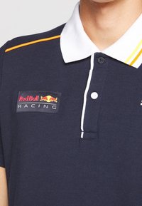 Polo shirt marine avec un col blanc, des rayures orange et jaune, et un logo Red Bull Racing sur la poitrine gauche. Tissu texturé, trois boutons.