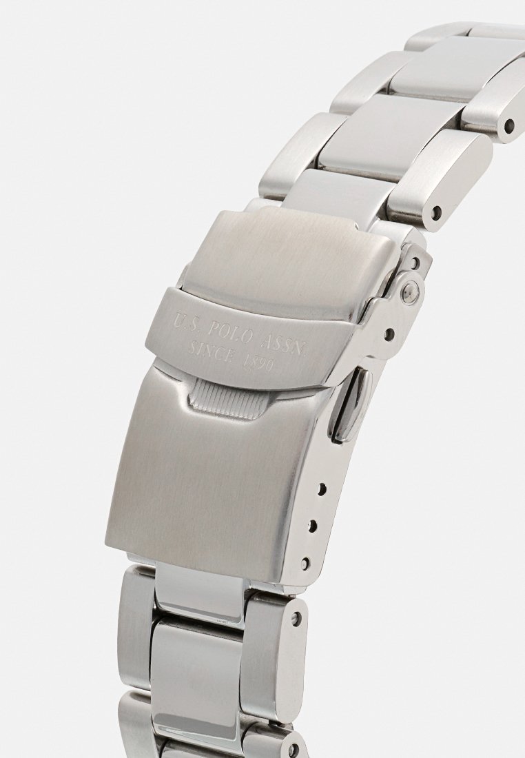 Montre en acier inoxydable avec une finition polie. Elle présente un bracelet segmenté, une fermeture par boucle et un marquage gravé sur la boucle.