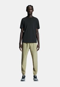 Sort kortærmet t-shirt parret med lyse khaki sweatpants og grå fritidssko, stående mod en ensfarvet grå baggrund.