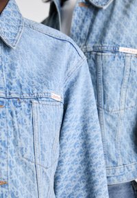Chaqueta de mezclilla azul claro con un patrón integral, cuello con puntas, bolsillos en el pecho y una etiqueta con el logo de "Calvin Klein" en la parte izquierda.