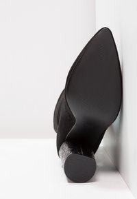 Bota negra de tacón alto con punta afilada, hecha de ante, que presenta un tacón de bloque de pelo negro texturizado y una suela de goma estriada.