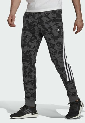 Uomo che indossa pantaloni da ginnastica Adidas mimetici neri e grigio scuro con strisce laterali bianche e scarpe da corsa nere.