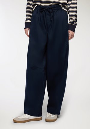 Pantaloni a gamba larga blu navy con vita elasticizzata e cordoncino; texture liscia, tasche laterali, abbinati a sneakers bianche e beige.