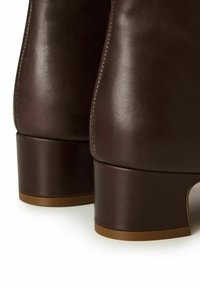 Brązowe skórzane botki z gładką fakturą, zaokrąglonym obcasem i kontrastującą jasnobrązową podeszwą. Charakteryzują się minimalnymi przeszyciami i elegancką sylwetką.