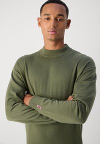 Pulover verde măsliniu cu un guler înalt și mâneci lungi. Manșeta are un mic accent cu logo roz brodat pe mânecă.