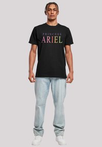 Čierne bavlnené tričko s nápisom "PRINCESS ARIEL" v gradientových farbách. Model má na sebe svetlomodré džínsy a biele tenisky.