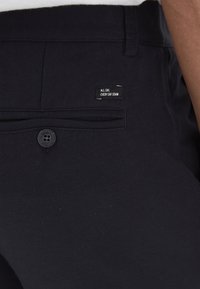 Pantaloni in denim nero con chiusura a bottone, una tasca posteriore con etichetta che recita "DENIM TUTTI I GIORNI, OGNI GIORNO" e una texture liscia.