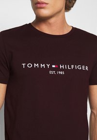 Burgundy cotton t-paita, jossa on pyöreä kaula-aukko, lyhyet hihat ja valkoinen brodeerattu logo punaisilla ja sinisillä yksityiskohdilla.
