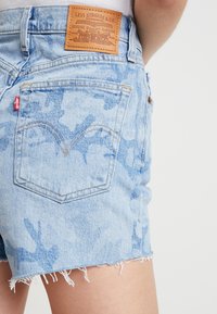 Light blue denim shorts with a faded camo pattern, frayed hem, and a leather patch at the back waistband with branding details.