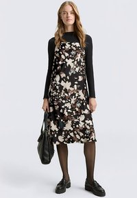 Zwarte bloemenjurk met bruine en crèmekleurige patronen, gladde stof, knielengte ontwerp, gedragen over een zwarte top met lange mouwen, gecombineerd met stevige schoenen.