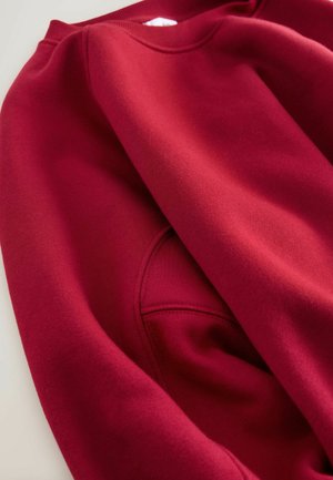 Hanorac roșu dintr-un material texturat, cu guler rotund și mâneci set-in, având cusături fine de-a lungul cusăturilor.