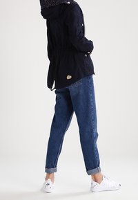 Person wearing a black jacket, blue cuffed jeans, and white Nike sneakers standing with hands in jacket pockets against a plain background.