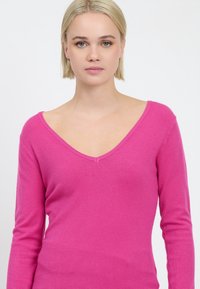 Maglione aderente di un vivace colore rosa, realizzato in materiale morbido. Presenta un ampio scollo a V e maniche lunghe, con una superficie liscia e dettagli minimi.