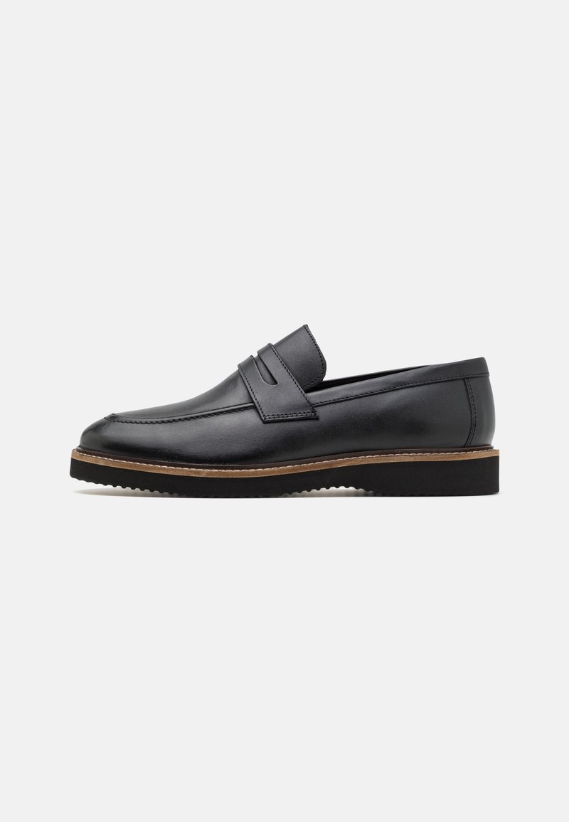 Chaussure en cuir noir à enfiler avec un bout rond, un double dessus à sangle et une semelle marron contrastante. Fini texturé avec une apparence lisse.