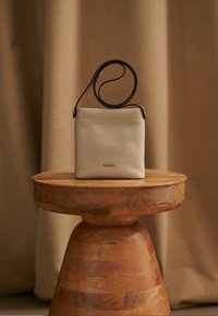 Lyse beige læder skuldertaske med tekstureret finish, med en kort sort rem, vist på en træpedestal.